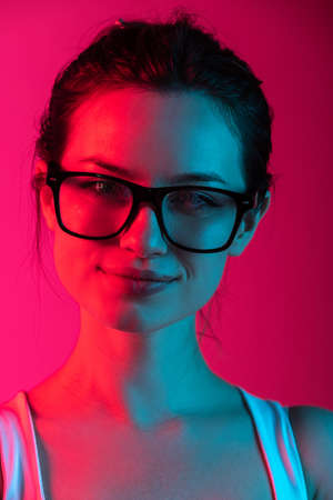 Close-up cropped portrait of young beautiful girl in glasses on gradient pink red background in neon lightsの写真素材