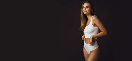 Cropped horizontal portrait of youn beautiful caucasian woman in white underwear on dark studio backgroundの写真素材
