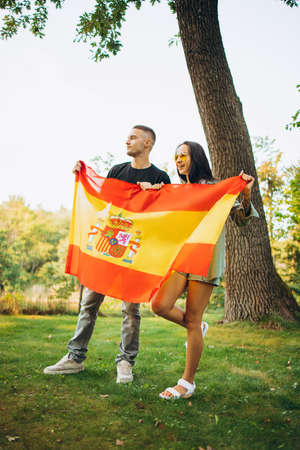 Full-length portrait of two young football fans, man and woman, holding flag of Spain, isolated on park backgroudndの写真素材