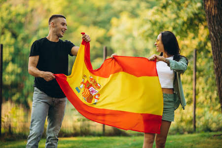 Portrait of two young football fans, man and woman, in a jump holding flag of Spain, isolated on park backgroudndの写真素材