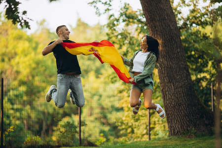 Portrait of two young football fans, man and woman, in a jump holding flag of Spain, isolated on park backgroudndの写真素材