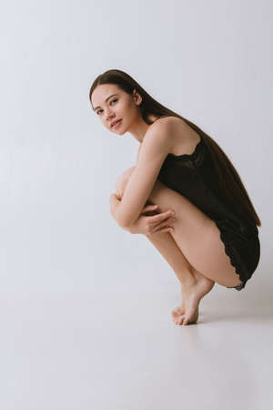 Full-lengths portrait of beautiful girl in black silk pajama sitting on grey studio wallpaperの写真素材