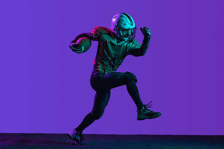 Full-length portrait of professional aerican football player in a run, training isolated over purple...