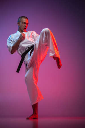 Full-length portrait of caucasian karate, judo, taekwondo sportsman in white kimono training isolated on gradient pink purple backgound in neon lightの写真素材