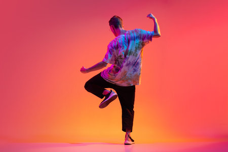 Back view of young stylish man dancing hip-hop isolated over colorful gradient background in neon...