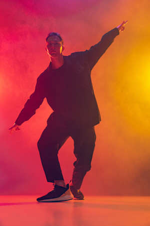 Young Caucasian man contemporary dancer in stylish black clothes in movement isolated over gradient pink yellow background in neon light in smokeの写真素材