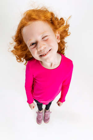 Portrait of little cute redheaded preschool girl in pink sweater posing isolated over white studio background. Winkingの写真素材