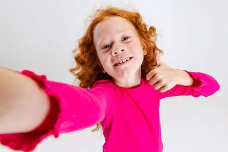 Portrait of little Caucasian girl, child posing and making selfie isolated over gray studio...