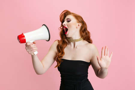 Cropped portrait of beautiful woman with retro hairstyle singing in megaphone isolated over pink backgroundの写真素材