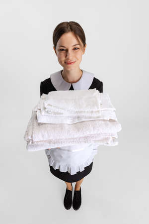 Top view portrait of young beautiful Caucasian housekeeper with stuck of fresh towels in uniform isolated over white backgroundの写真素材