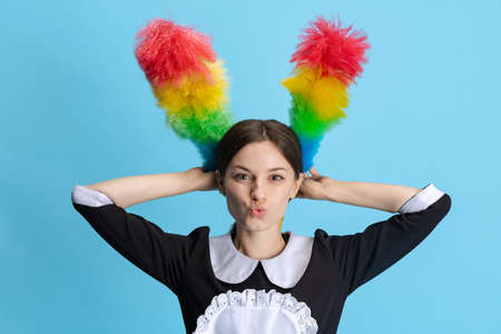 Funny and beautiful young woman, chambermaid in uniform holding pipidastre like bunny ears isolated over blue backgroundの写真素材