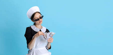 Bossy young woman in chambermaid uniform and stylish sunglasses counting money isolated over blue backgroundの写真素材