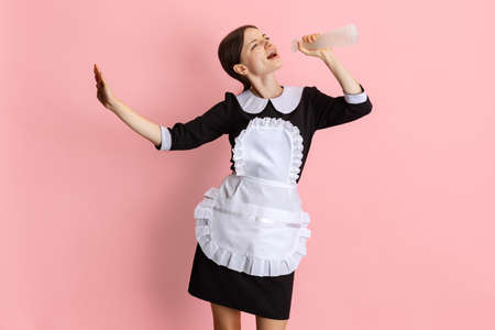Portrait of young beautiful hotel chambermaid in black uniform singing in cleaning spray bottle isolated over pink background.の写真素材