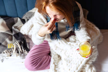 Cropped portrait of young sick girl sitting on sofa at home and drinking medicine. Sneezingの写真素材
