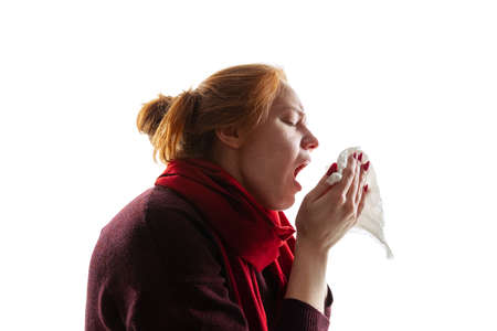 Portrait of young girl having a cold and flu, sneezing into napkin isolated over white background. Seasonal illness, allergyの写真素材