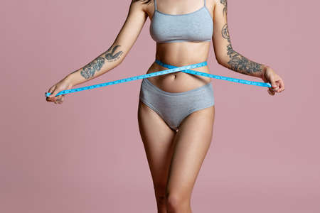 Cropped portrait of beautiful female body in gray inner wear, measuring waist isolated over pink background. Body positivityの写真素材