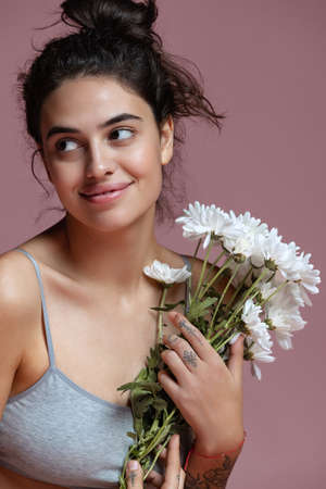 Cropped portrait of beautiful young woman in gray inner wear holding flowers isolated over pink...