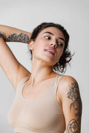 Cropped portrait of young beautiful woman holding hair in ponytail with natural makeup in beige inner wear isolated over gray backgroundの写真素材