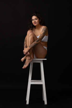 Full-length portrait of beautiful slim girl in white cotton underwear posing on chair isolated over dark backgroundの写真素材