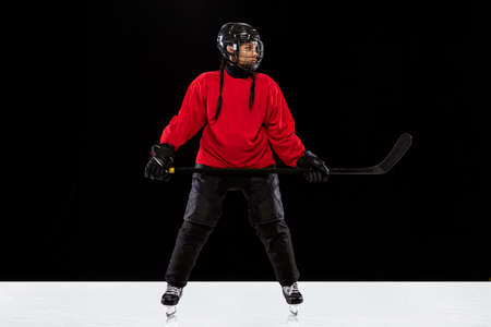 Full-length portrait of woman, professional hockey player in special protective uniform with helmet and hockey stick isolated over black background.の写真素材