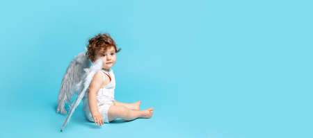 Side view portrait of cute toddler boy, baby in white romper with angel wings isolated over blue...