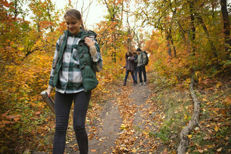 Young girl walking in the forest, holding thermos with hot tea. Group of friends doing autumn hiking togetherの写真素材