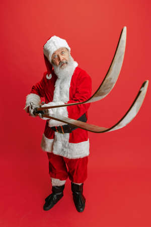 Full-length portrait of senior man wearing Santa Claus costume, holding skis isolated over red background. Winter holiday seasonの写真素材