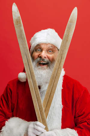 Cropped portrait of joyful senior man wearing Santa Claus costume, holding skis isolated over red background. Winter holiday seasonの写真素材