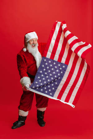 Full-length portrait of senior man wearing Santa Claus costume, holding american flag isolated over red backgroundの写真素材