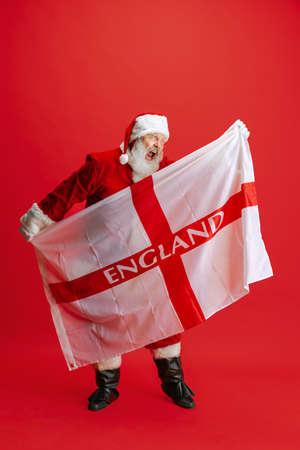 Full-length portrait of senior man wearing Santa Claus costume, holding England flag isolated over red backgroundの写真素材