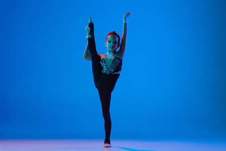 Full-length portrait of little girl, professional rhythmic gymnast standing in twine position with ball isolated over blue background in neonの写真素材