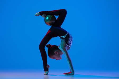 Full-length portrait of little girl, professional rhythmic gymnast training, performing isolated over blue background in neonの写真素材