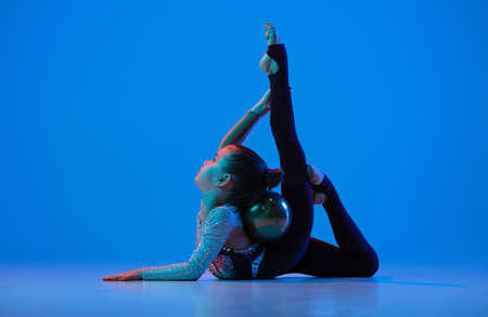 Full-length portrait of little girl, professional rhythmic gymnast training, performing isolated over blue background in neonの写真素材