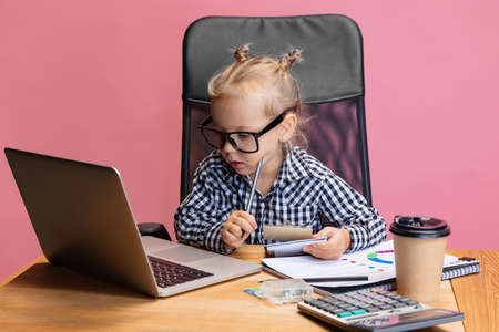 Cropped portrait of cute little girl sitting in front of laptop and making notes isolated over pink...