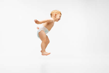 Full-length portrait of cute little boy, playful child in diaper jumping isolated over white...