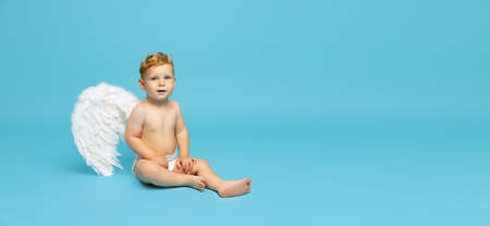 Full-length portrait of little boy, child in diaper with angel wings calmly sitting isolated over blue background. Flyerの写真素材
