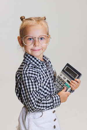 Cropped side view portrait of little cute girl, child in skirt and checkered shirt with calculator isolated over gray backgroundの写真素材