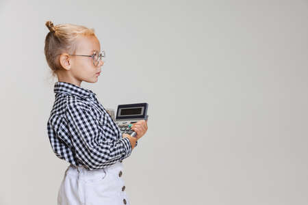 Cropped side view portrait of little cute girl, child in skirt and checkered shirt with calculator isolated over gray backgroundの写真素材