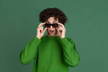 Cropped portrait of young man in green sweater and stylish sunglasses isolated over dark green background. Fashionable youthの写真素材