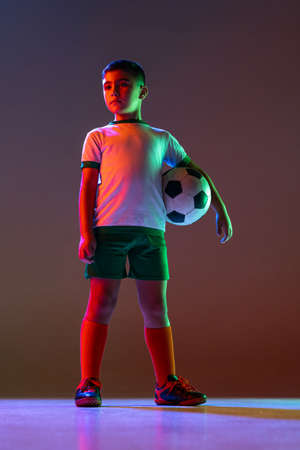 Full-length portrait of boy, child, football player in uniform with ball posing isolated over gradient background in neon light.の写真素材