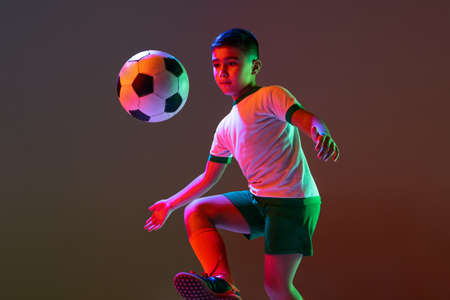 Cropped portrait of boy, child, football player training with ball isolated over gradient background in neon light.の写真素材