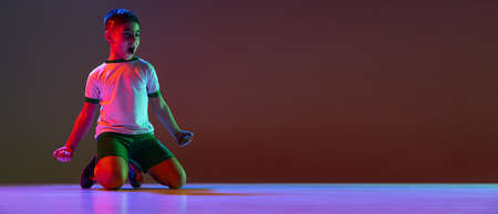 Full-length portrait of boy, child, football player sitting in winning pose isolated over gradient...
