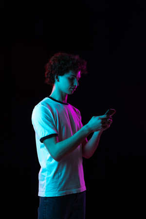 Cropped portrait of young man in white T-shirt with phone isolated over black background in neon lightsの写真素材