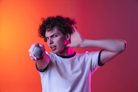 Cropped portrait of young man in white T-shirt directing microphone on camera isolated over gradient pink orange background in neon lightsの写真素材