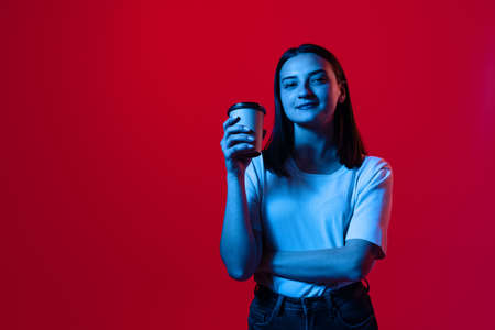 Cropped portrait of young beautiful woman in white T-shirt with coffee cup isolated over red...