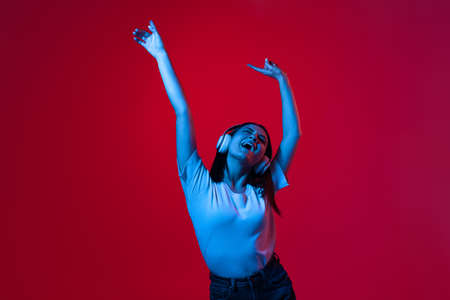 Cropped portrait of young beautiful woman in white T-shirt listening to music in headphones and dancing isolated over red background in neon lightsの写真素材