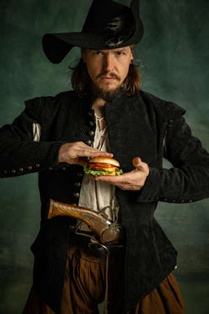 Portrait of one brutal man, medeival pirate squeezing burger isolated over dark background.の写真素材