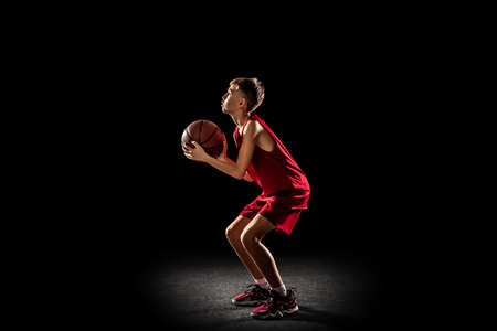 Full-length portrait of teen boy, playing, training basketball isolated over black background....