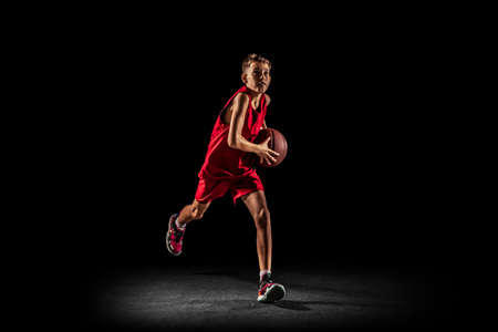 Full-length portrait of teen boy, basketball player in motion, training, running with ball isolated over black background. Goal scoringの写真素材