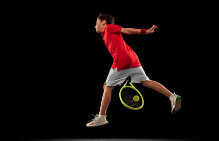 Portrait of sportive young boy, tennis player in unifrom, training isolated over black backgound. In...